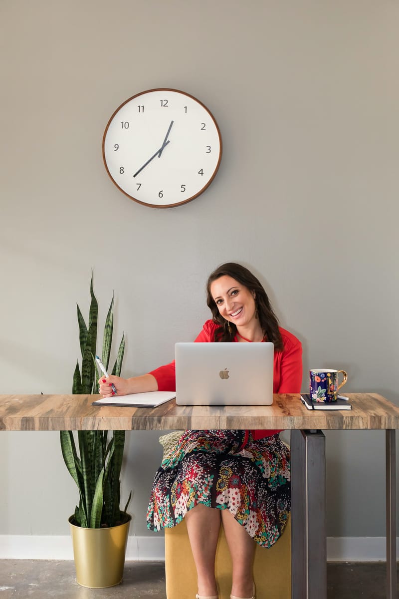 Lauren Goldstein working at her desk with a laptop and notebook, smiling, beneath a wall clock